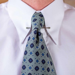 May include: A close-up of a white dress shirt with a patterned tie and tie bar. The tie features a repeating pattern of blue diamonds and circles on a light green background. The tie bar is a thin, silver-colored metal bar.