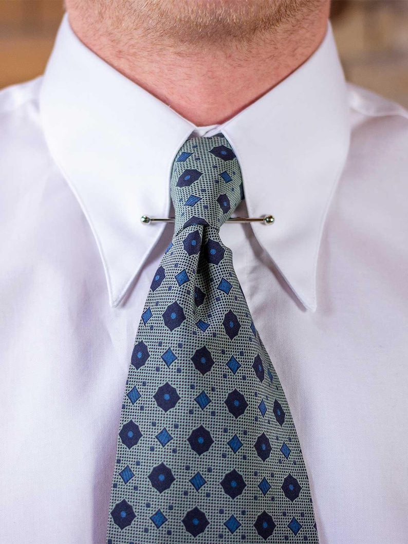 May include: A close-up of a white dress shirt with a patterned tie and tie bar. The tie features a repeating pattern of blue diamonds and circles on a light green background. The tie bar is a thin, silver-colored metal bar.