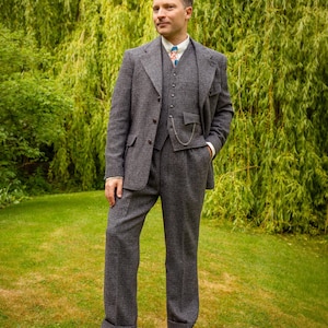 May include: A three-piece suit in a dark grey and white micro-check pattern, consisting of a single-breasted jacket, a matching waistcoat with a silver pocket watch chain, and trousers with cuffs, worn with a white shirt and two-tone brown shoes.