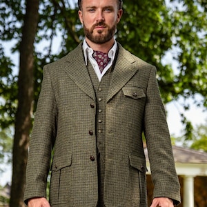 May include: A three-piece tweed suit in shades of olive green and brown. The suit includes a jacket with two front pockets, a vest, and matching trousers. A white collared shirt and patterned ascot complete the ensemble. The suit is tailored and appears to be made of a textured fabric.