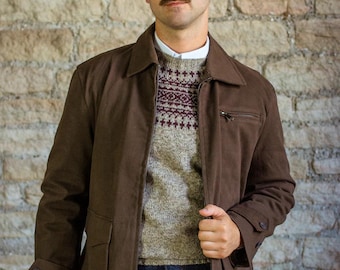Può includere: Giacca marrone con collo e chiusura a zip. La giacca è indossata sopra un maglione fantasia e una camicia bianca con colletto. La persona indossa jeans blu. Lo sfondo è un muro di pietra.