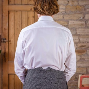 May include: A white dress shirt with long sleeves and a button-down collar. The shirt is paired with gray tweed trousers. The person is standing in front of a wooden door and a brick wall.