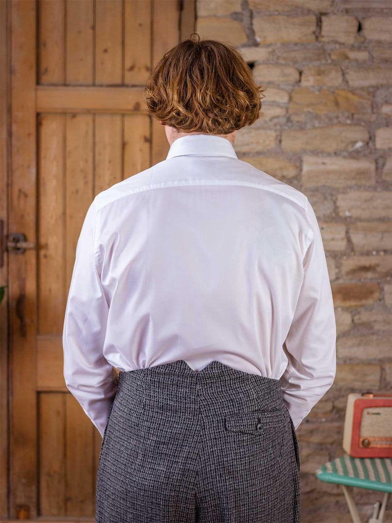 May include: A white dress shirt with long sleeves and a button-down collar. The shirt is paired with gray tweed trousers. The person is standing in front of a wooden door and a brick wall.