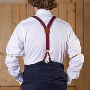 May include: A white long-sleeved button-down shirt paired with dark charcoal trousers and burgundy braces featuring light grey trim and brown leather accents. The shirt sleeves are held in place by metallic gold sleeve garters.