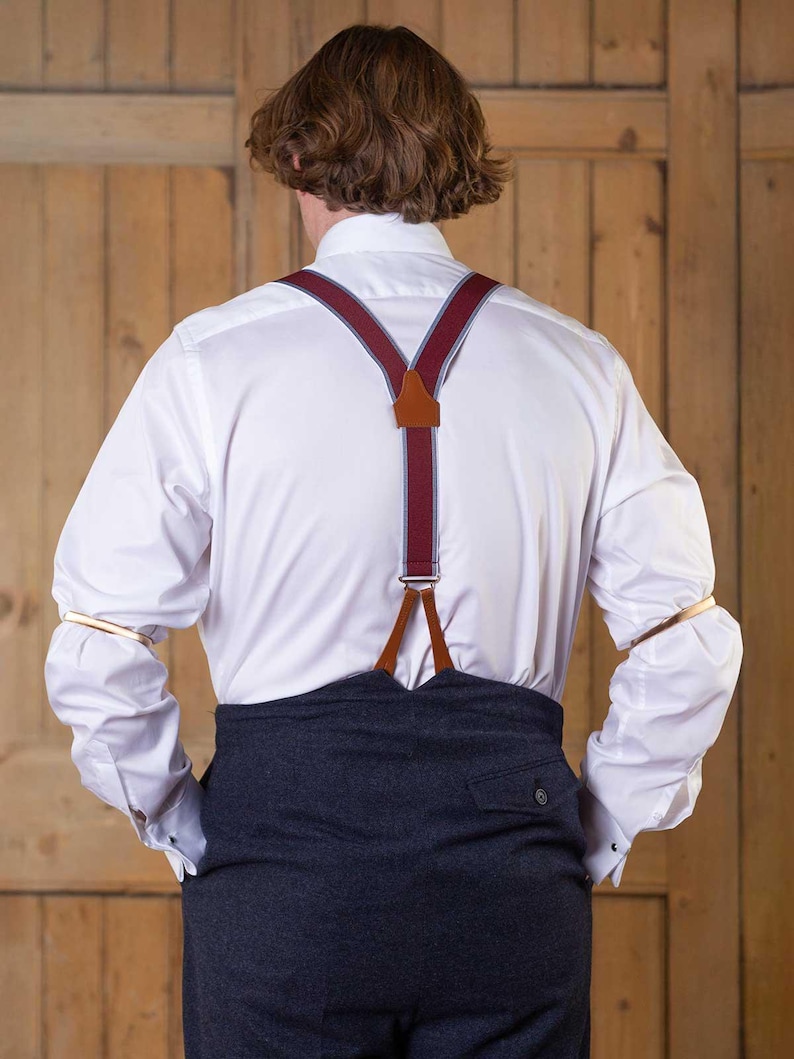 May include: A white long-sleeved button-down shirt paired with dark charcoal trousers and burgundy braces featuring light grey trim and brown leather accents. The shirt sleeves are held in place by metallic gold sleeve garters.