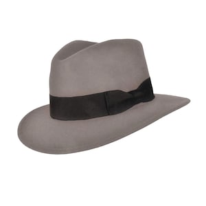 Könnte beinhalten: Ein hellgrauer Fedora-Hut mit einer eingedrückten Krone und einer breiten, strukturierten Krempe. Der Hut ist mit einem einfarbigen schwarzen Ripsband verziert, das die Basis der Krone umschließt und einen Kontrast zum grauen Filz bildet.