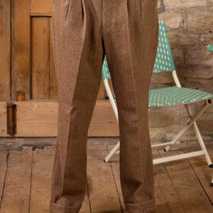 Peut inclure: Pantalon en tweed marron à plis et ourlets retroussés. Le pantalon fait partie d'un costume, avec un gilet assorti visible. La personne porte des chaussures en cuir marron. L'arrière-plan comprend une porte en bois et une chaise vert clair.