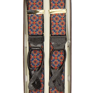 May include: A pair of patterned suspenders in a box. The suspenders feature a repeating design of red and gold interlocking circles on a navy blue background. They have black leather attachments and gold-colored metal clasps.