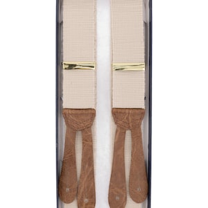 Può includere: Bretelle beige con chiusure in metallo color oro e estremità in pelle marrone, presentate in una scatola. Le bretelle sono realizzate in tessuto intrecciato.