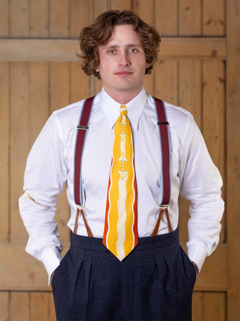 May include: A crisp white long-sleeved button-down shirt paired with dark charcoal pleated trousers. The outfit features a vibrant yellow necktie with a white geometric pattern and burgundy braces with silver hardware and brown leather tabs.