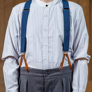 Könnte beinhalten: Ein weißes Hemd mit Knöpfen und blau-weißen Streifen, getragen mit einer dunkelgrauen Hose und blauen Hosenträgern mit braunen Lederakzenten.