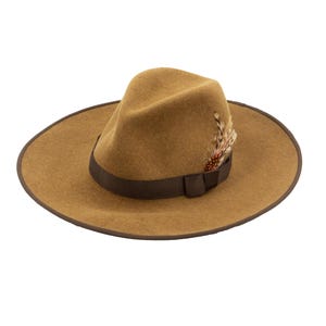 May include: A tan felt fedora hat featuring a wide brim with a dark brown edge trim. The hat includes a dark brown ribbon band around the crown and is accented with a small cluster of brown and white feathers tucked into the side of the band.