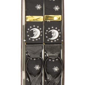 May include: Black suspenders with a celestial design featuring white crescent moons, stars, and suns. The suspenders have gold-colored metal clasps and black leather loops. The suspenders are packaged in a box.