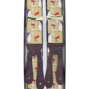 May include: A pair of suspenders with a repeating pattern of white figures holding red flags on a tan background. The suspenders have brown leather ends and gold-colored metal clasps. The suspenders are displayed in a dark brown box.