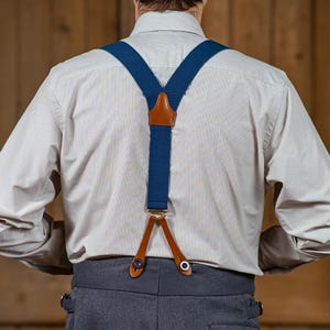 May include: A pair of blue suspenders with brown leather accents. The suspenders are attached to a white button-down shirt and gray pants.