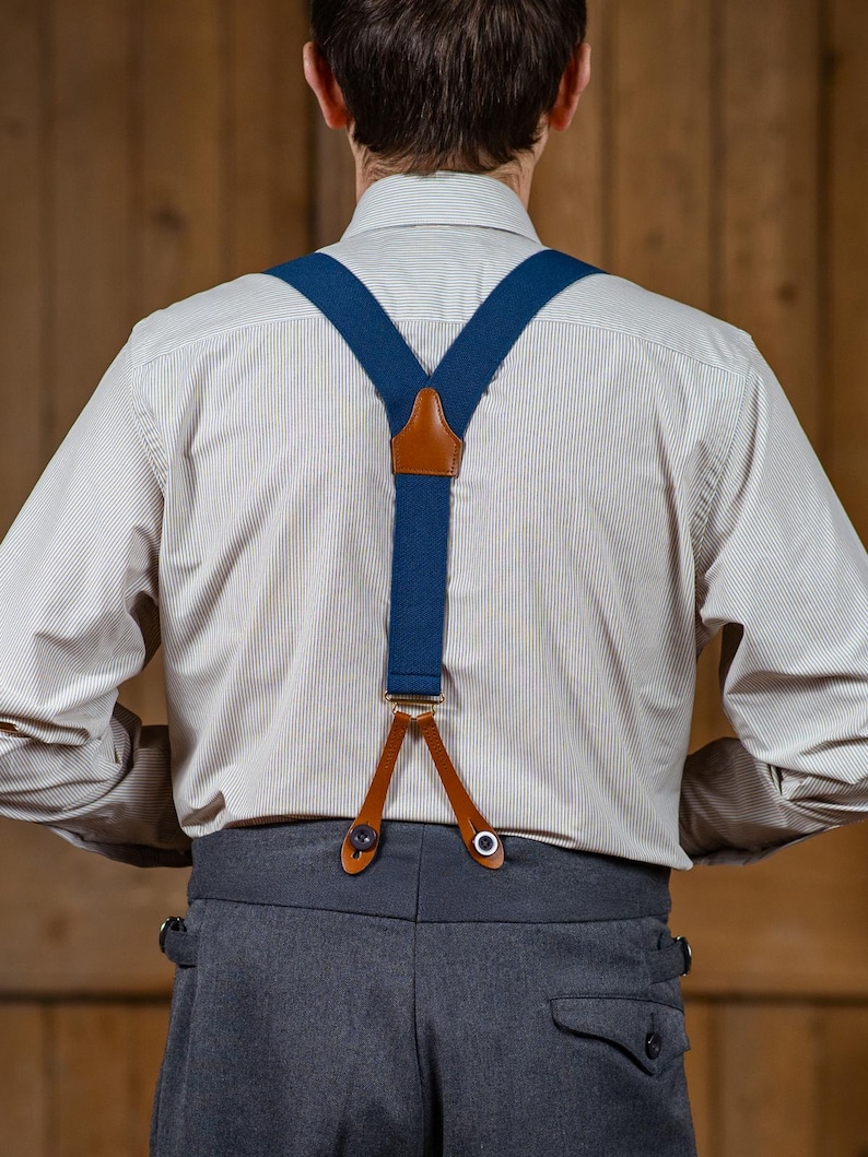 May include: A pair of blue suspenders with brown leather accents. The suspenders are attached to a white button-down shirt and gray pants.