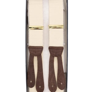 Può includere: Bretelle color crema con chiusure dorate ed estremità in pelle marrone scuro, presentate in una scatola bianca e nera. Le bretelle hanno una trama strutturata e sono progettate per essere indossate con pantaloni.