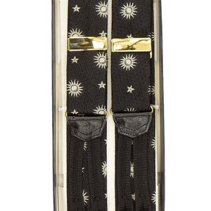 May include: Black suspenders with a repeating pattern of white suns and stars, presented in a box. The suspenders have gold-colored metal clasps and black leather-like tabs.