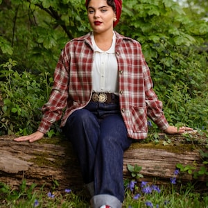 May include: A person wearing a red headscarf, white blouse, and a red and brown plaid jacket. They are wearing dark blue jeans and red and white shoes. A gold belt with a decorative buckle is visible. The person is sitting on a log in a natural setting.
