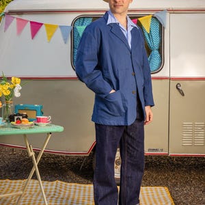 May include: A blue blazer with two front pockets, a light blue and white striped collared shirt, dark blue denim jeans with cuffed hems, brown leather shoes, and a straw fedora hat with a blue, white, and black striped band.