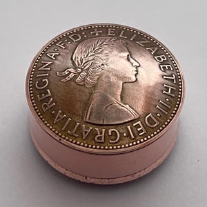 May include: A circular box in a copper hue, with a coin-like top. The lid displays a profile and the words "REGINA F.D. FELIZABETH II DEI GRATIA". The box has a simple, cylindrical form, suitable for small items.