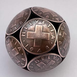 May include: A spherical sculpture constructed from multiple metallic coins, each featuring a central cross emblem, the text HELVETIA, and the year 1948. The coins are arranged to form a rounded, ball-like shape with a dark, weathered patina finish.