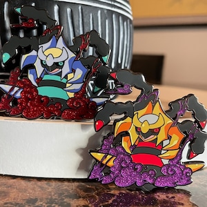 May include: Two enamel pins featuring a stylized creature design. One pin has a blue, white, and teal color scheme with red glitter accents. The other pin has a yellow, orange, and red color scheme with purple glitter accents.