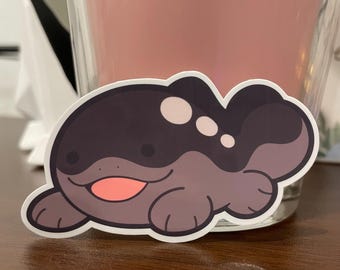 May include: A sticker of a cartoon creature with a dark gray body, white spots, and a pink mouth. The creature has a rounded shape and is lying on its belly.