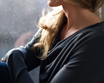 May include: A woman with long blonde hair is wearing a dark gray, cowl neck top. She is sitting by a window and looking out.