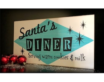 May include: A rectangular wooden sign with the words "Santa's DINER" in black letters on a white background. A turquoise diamond shape is behind the word "DINER". The sign also says "Serving warm cookies & milk". Red Christmas ornaments are in front of the sign.