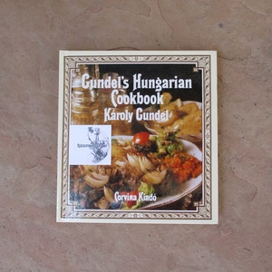 May include: A cookbook titled "Gundel's Hungarian Cookbook" by Károly Gundel. The cover features a photo of a Hungarian dish with meat, vegetables, and a side of potatoes. The book has a decorative border and the publisher's name, "Corvina Kiadó."
