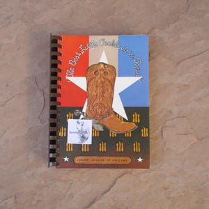 May include: A spiral-bound cookbook titled "The Best Little Cookbook in Texas" featuring a brown cowboy boot on a Texas flag design. The cover includes the text "Junior League of Abilene" and a small sticker.