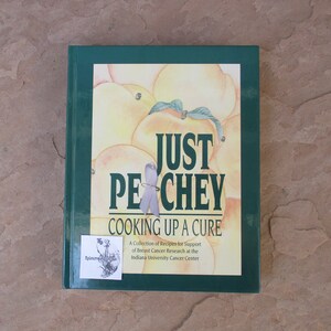 May include: A hardcover cookbook titled "Just Pechey Cooking Up a Cure" with a green spine and cover. The cover features a watercolor illustration of peaches and a light purple ribbon. A sticker with text is on the lower left.