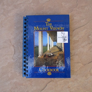 May include: A blue spiral-bound cookbook titled "The Mount Vernon Cookbook." The cover features a color image of a porch with white columns, chairs, and a table set for tea. The cookbook has a small sticker on the front.