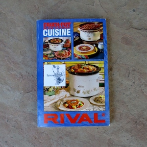 May include: A vintage cookbook titled "Crock-Pot Slow Cooker Cuisine" by Rival. The cover is blue with red and white text and features images of slow-cooked meals. The cookbook is on a stone surface.