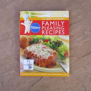 May include: A cookbook titled "Pillsbury Family Pleasing Recipes" with the Pillsbury Doughboy logo. The cover features a photo of a lasagna dish with salad. The text reads "170 Super-Fast and Easy Recipes That Everyone Will Love."