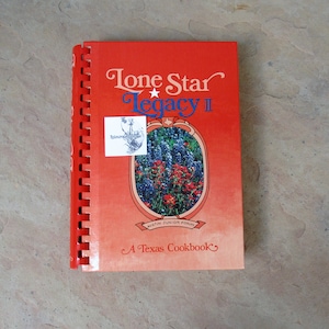 May include: A red spiral-bound cookbook titled "Lone Star Legacy II: A Texas Cookbook." The cover features a floral illustration within an oval frame and the text "Austin Junior Forum."