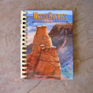May include: A spiral-bound cookbook titled "West of the Rockies" with a landscape illustration. The cover features a large rock formation in shades of orange and brown against a blue sky. Text includes "Recipes from Campfire to Candlelight".
