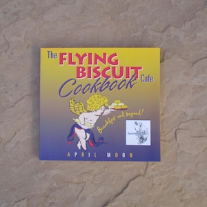 May include: A cookbook titled "The Flying Biscuit Cafe Cookbook" with a yellow, blue, and purple cover. The cover features an illustration of a cherub with wings holding biscuits. The author's name, April Moon, is at the bottom.