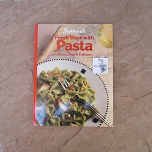 May include: A cookbook titled "Sunset Fresh Ways with Pasta" featuring a photo of pasta on a plate. The cover has the text "Healthy, Easy & Delicious!" and a small sticker. The book is on a brown stone surface.