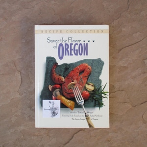 May include: A cookbook titled "Savor the Flavor of Oregon" with a white cover. The cover features an illustration of food, including a piece of salmon, mushrooms, and tomatoes. The book is part of a recipe collection.