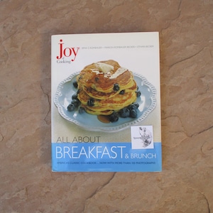May include: A cookbook titled "Joy of Cooking" with the subtitle "All About Breakfast & Brunch." The cover features a stack of pancakes with blueberries and butter on a light blue plate. The book is a classic American cookbook.