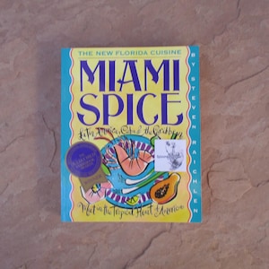 May include: A cookbook titled "Miami Spice" with a colorful cover featuring illustrations of tropical fruits and seafood. The book has a blue border and yellow background with the title in large, bold letters. The book is a winner of the Julia Child Cookbook Award.
