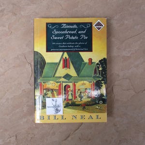 May include: A hardcover cookbook titled Biscuits, Spoonbread, and Sweet Potato Pie by Bill Neal. The yellow cover features an illustration of a house with a green roof and red shutters, with people depicted in the front yard and on the porch.