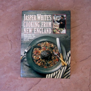 May include: A cookbook titled "Jasper White's Cooking From New England" with a photo of a seafood dish on the cover. The cover also features a smaller image of a person at a market. The book is on a textured surface.
