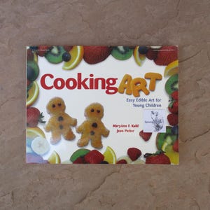 May include: A softcover book titled Cooking Art: Easy Edible Art for Young Children by MaryAnn F. Kohl and Jean Potter. The white cover features a border of colorful fruit slices and two small golden-brown gingerbread-style figures in the center.