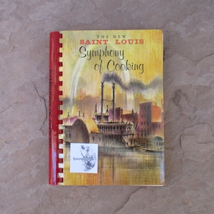 May include: A vintage cookbook titled "The New Saint Louis Symphony of Cooking." The cover features an illustration of a steamboat on a river with buildings and a yellow and orange background. The spine is red.