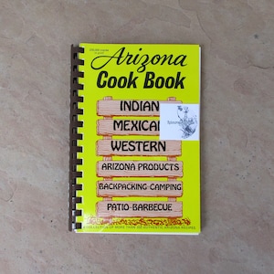 May include: A spiral-bound cookbook titled "Arizona Cook Book" with a bright yellow cover. The cover features the title in large, black script and lists recipe categories like Indian, Mexican, and Western. The book contains over 350 authentic Arizona recipes.