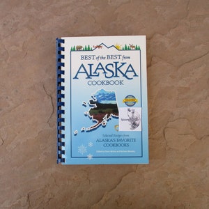 May include: A cookbook titled "BEST of the BEST from ALASKA COOKBOOK" with a blue spiral binding. The cover features an outline of Alaska, illustrations of mountains, trees, and wildlife, and the text "Selected Recipes from Alaska's Favorite Cookbooks."