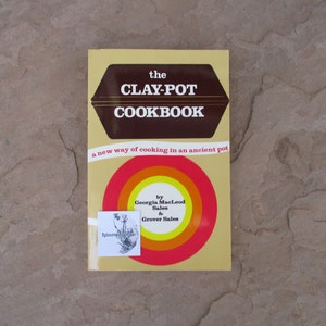 May include: A vintage cookbook titled "The Clay-Pot Cookbook" with a brown and gold cover. The cover features a brown hexagon with the title in white, and a circular design with red, orange, and yellow rings. The book promotes a new way of cooking.